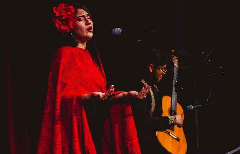 A female singer in a red shawl and flower hair accessory performs with a live acoustic guitarist.