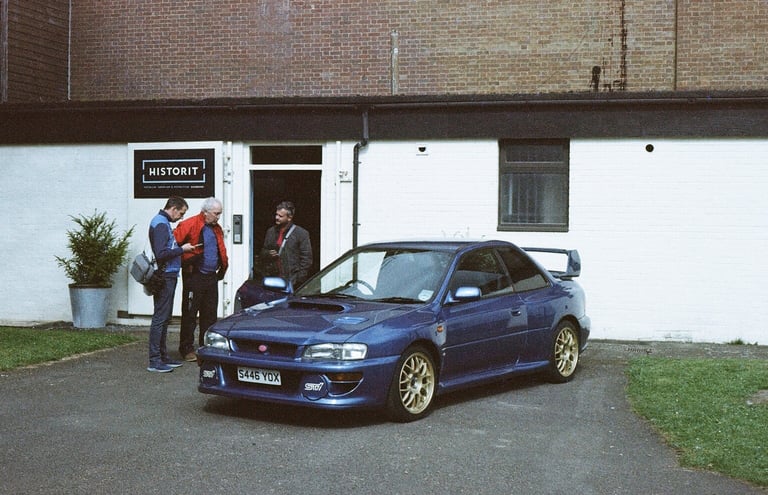 a classic Subaru Impreza parked in front a building with three guys looking at it