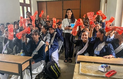 About 20 girls in classroom holding up napkin packs. with Pooja.