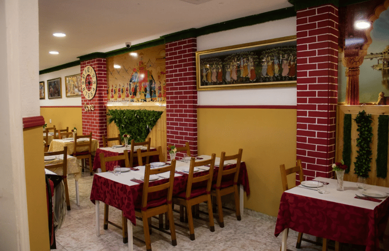 Indian restaurant interior with red patterned tablecloths, wooden chairs, and traditional murals on yellow walls.