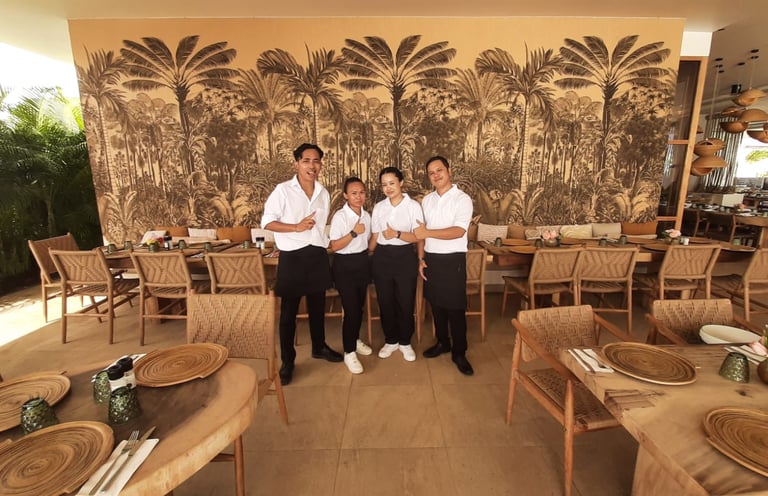 Restaurant service team posing in a tropical-themed dining room with palm tree wallpaper.