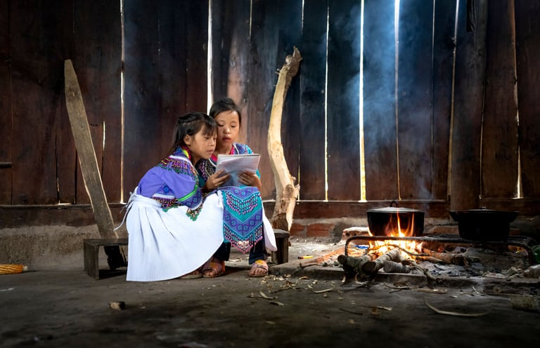 Two young girls in traditional attire reading together by an indoor cooking fire.
