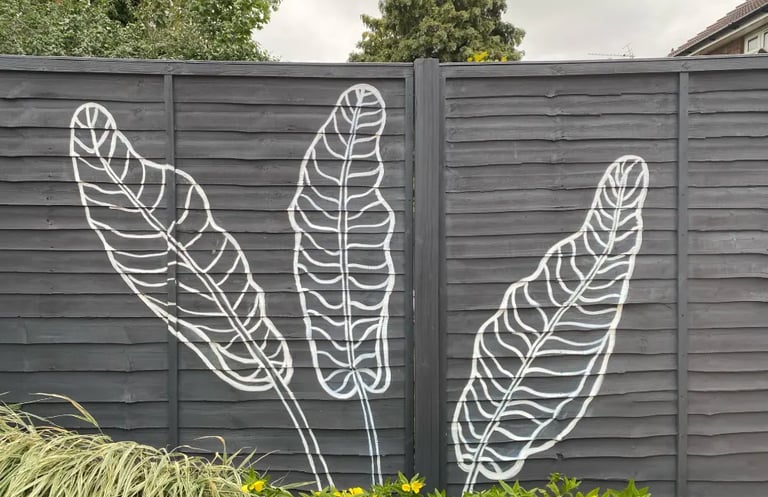 Three large banana leaves painted in a modern graphic style on a grey yard fence and flowers.