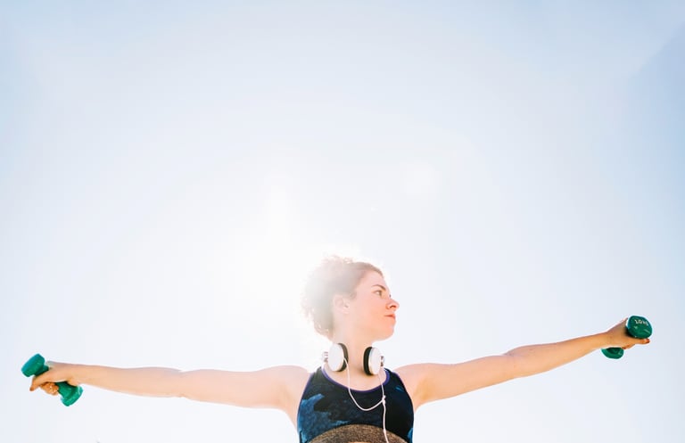 A woman exercising outdoors with green dumbbells and headphones during a sunny day workout.