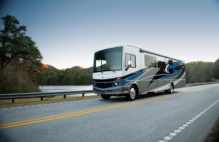 A luxury Class A Fleetwood motorhome drives on a scenic mountain road next to a calm lake.
