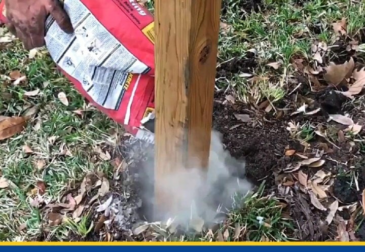 A person pouring fast-setting concrete mix from a bag into a hole to set a wooden fence post in the ground.