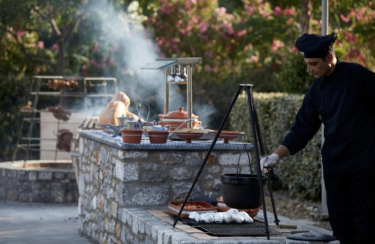 A professional chef cooking traditional Greek food over an outdoor stone grill with a hanging tripod pot.