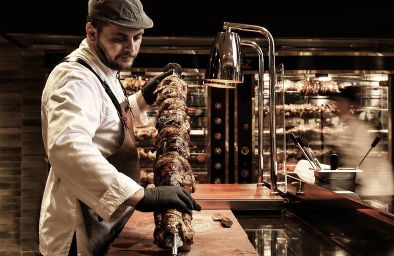 Professional chef preparing a vertical rotisserie spit of roasted meat in a restaurant kitchen.