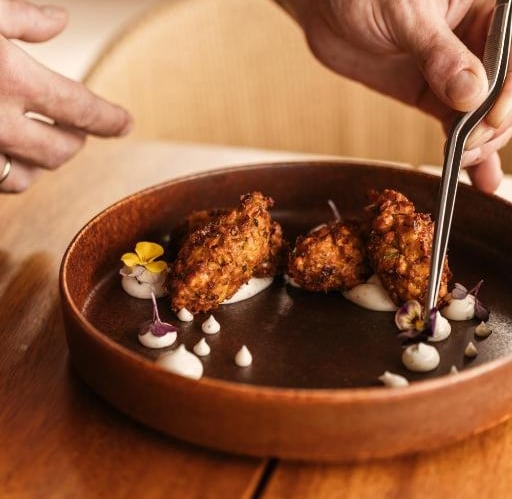 Chef plating gourmet vegetable fritters with edible flowers and cream sauce in a restaurant.