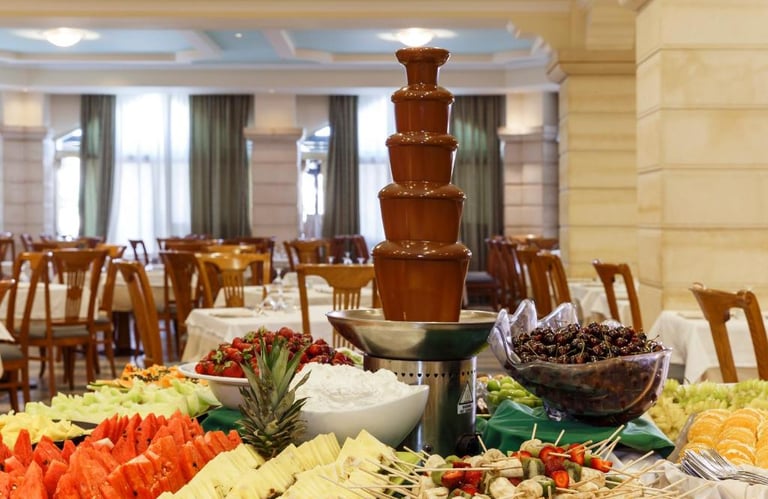 A tiered chocolate fountain on a buffet table surrounded by fresh fruit platters in a hotel restaurant.