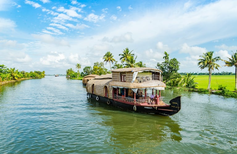 Houseboat cruising through Kerala's backwaters, surrounded by lush palm trees and vibrant greenery under a blue sky.