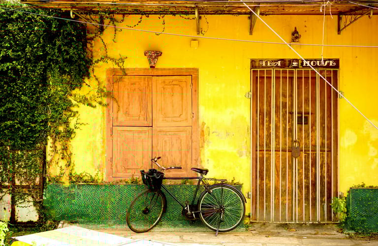 Bicycle parked in front of an old tea house with yellow walls and wooden doors, surrounded by lush green vines, Fort Kochi.
