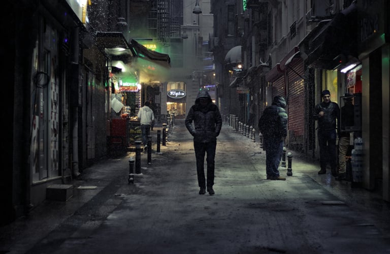Pessoa de casaco andando em um beco à noite, em bairro humilde, transmitindo solidão e introspecção.