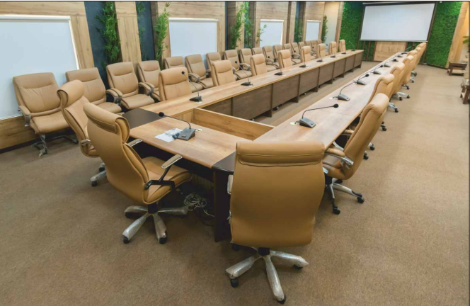Modern conference room with tan leather chairs and a long wooden U-shaped table with microphones.