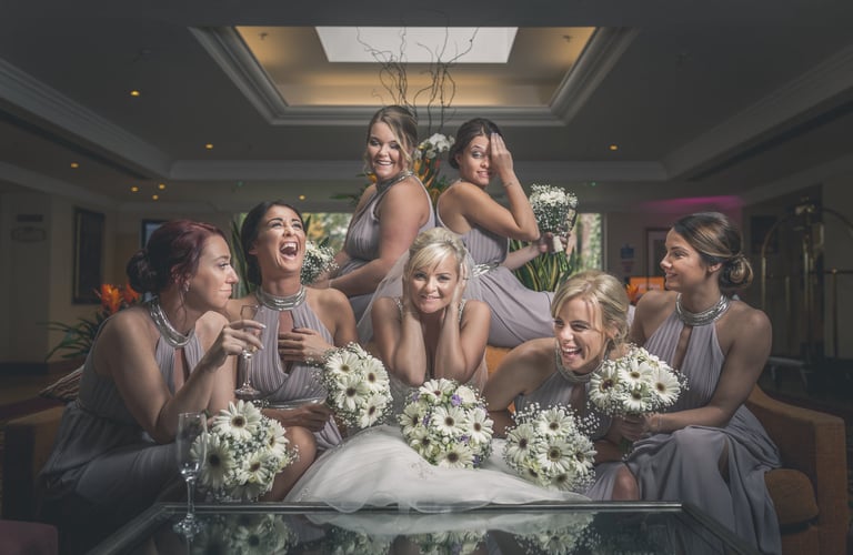 Smiling bride and bridesmaids in grey dresses holding white gerbera daisy bouquets at a wedding reception.