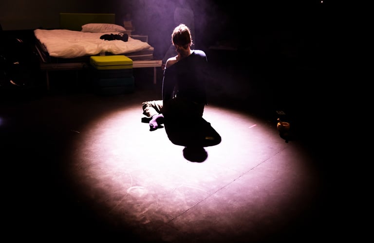 Tatum sits on the floor looking away from the audience, bathed in a spotlight facing their bed
