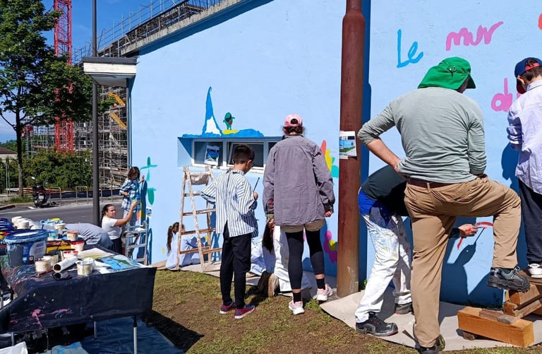 Gruppo di bambini e adulti dipinge insieme su muro azzurro durante laboratorio artistico Poliops.