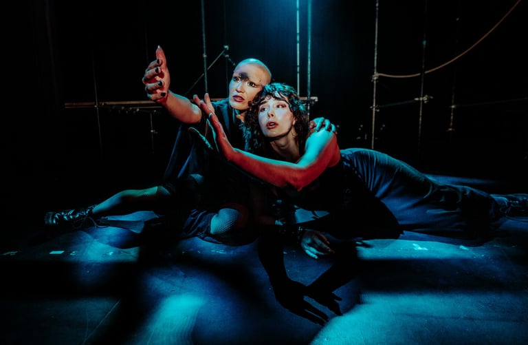 Ila and Coda, bathed in blue light and shadow reach their arms up together, their gazes meeting