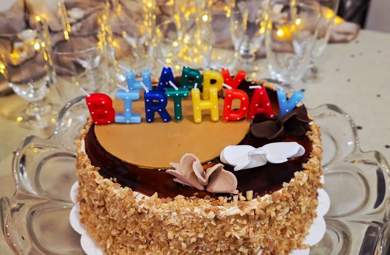 Chocolate birthday cake with nut topping and colorful Happy Birthday candles on a glass stand.