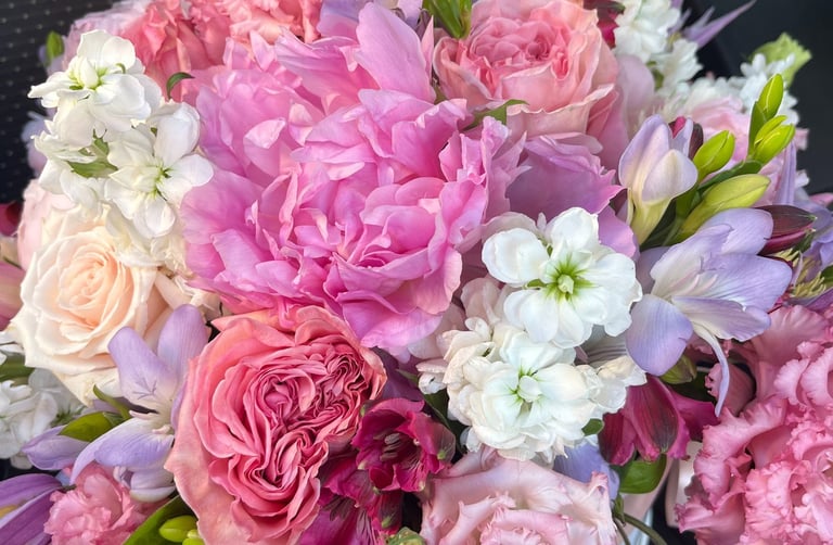 A vibrant pink and purple floral arrangement featuring peonies, roses, and white stock flowers.