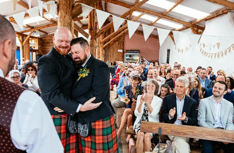 Grooms embrace during wedding ceremony in rustic barn at South Wales venue Ceridwen, with guests clapping and cheering