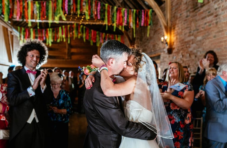 Bride and groom kiss during barn wedding ceremony in South Wales, with guests applauding under colourful ribbon decor