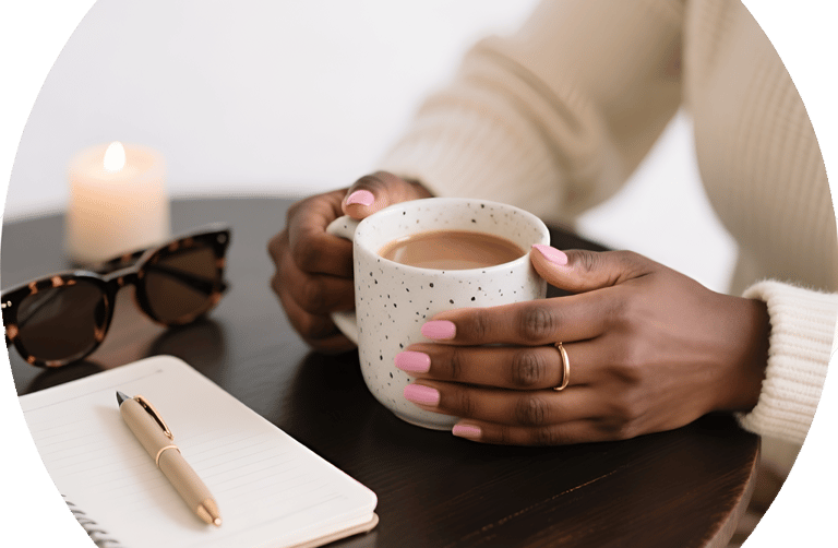 A woman holds a ceramic coffee mug on a table with a notebook, pen, and sunglasses.