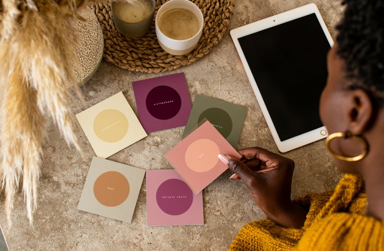 A woman holds colorful affirmation cards on a desk with a digital tablet and coffee.