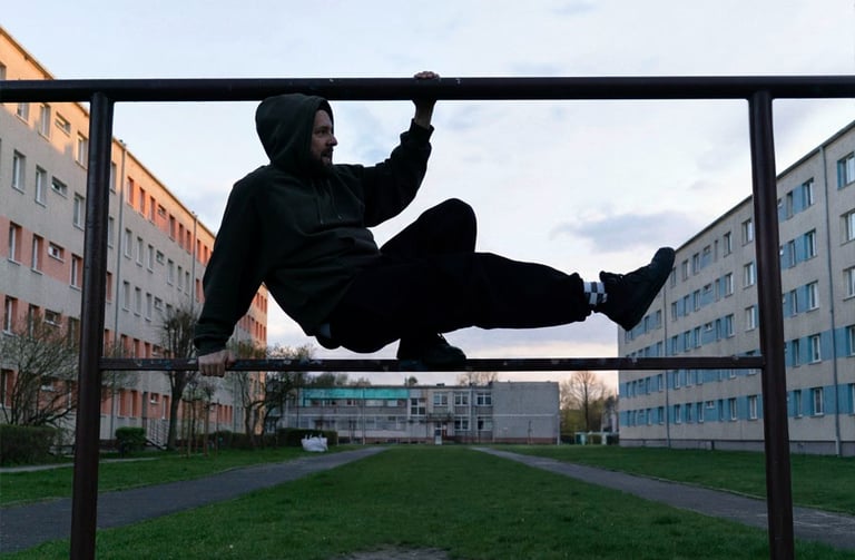 A man in a green hoodie performs parkour moves on a metal frame in an urban residential courtyard.