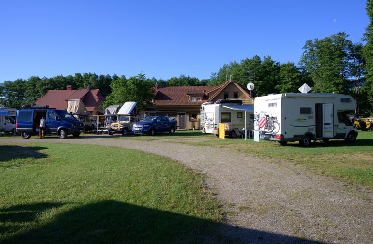 A sunny motorhome campsite with camper vans and RVs parked near a lodge in a grassy forest area.