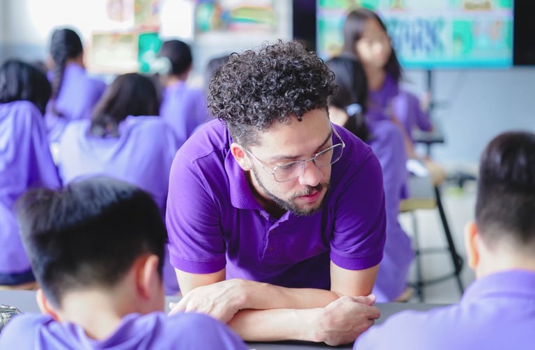 Male teacher leaned overlooking at a students work. In front of the teacher are two students leaned over doing their work.