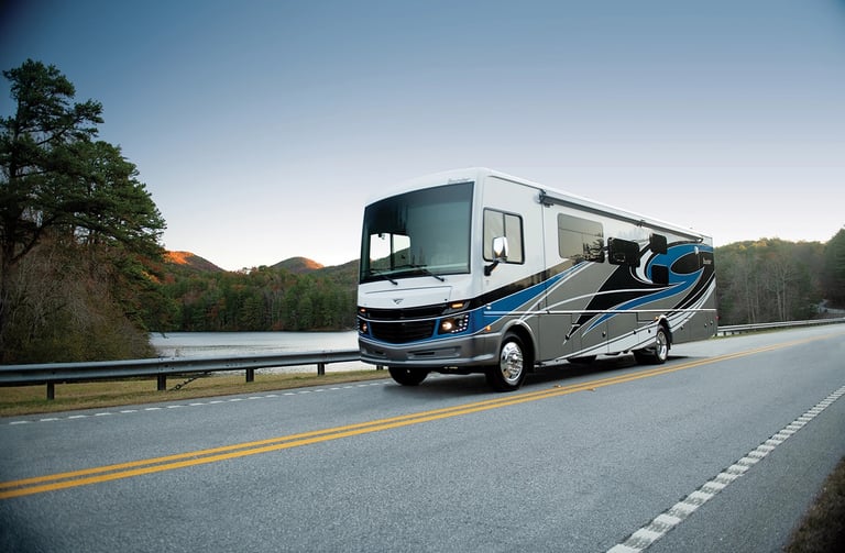 Modern Class A motorhome traveling on a scenic highway alongside a lake and autumn mountains.