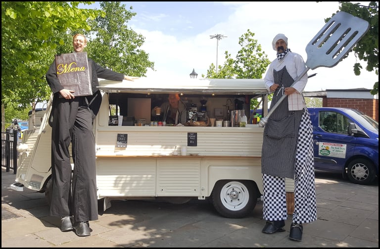 Two men on stilts, one holding a menu board and the other a giant spatula, stand beside a food truck selling Jamaican food.