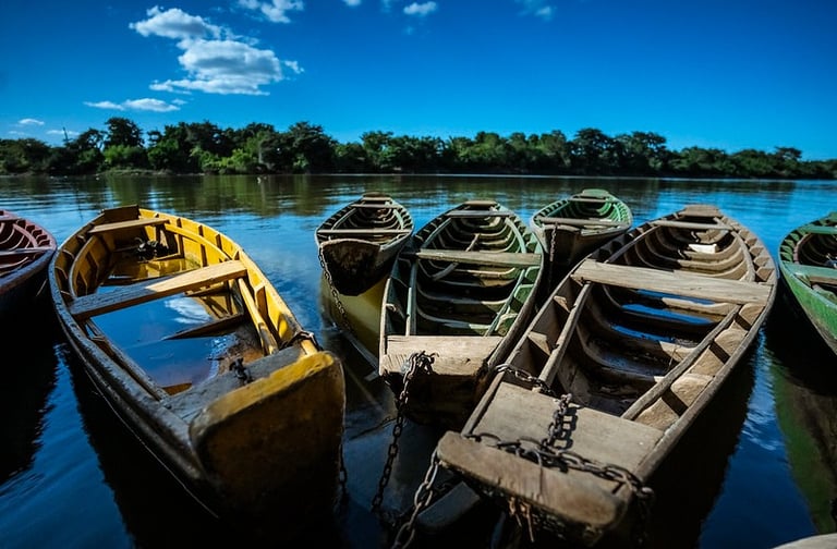 Rustic wooden rowboats anchored on a calm river under a clear blue sky with green trees.
