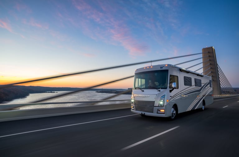 A Winnebago Class A motorhome traveling across a suspension bridge at sunset.