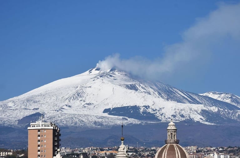 Panoramic view of snow-capped Mount Etna volcano emitting smoke above the skyline of Catania, Sicily.