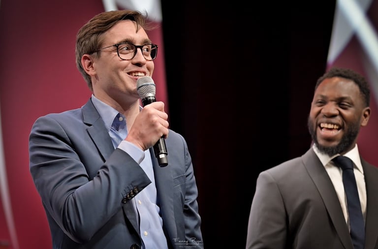 A smiling male speaker in a blue suit holding a microphone at a conference stage