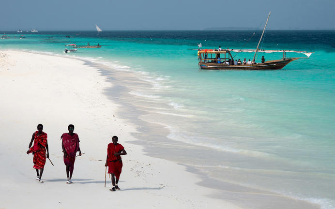 three people walking on a beach with boats in the background