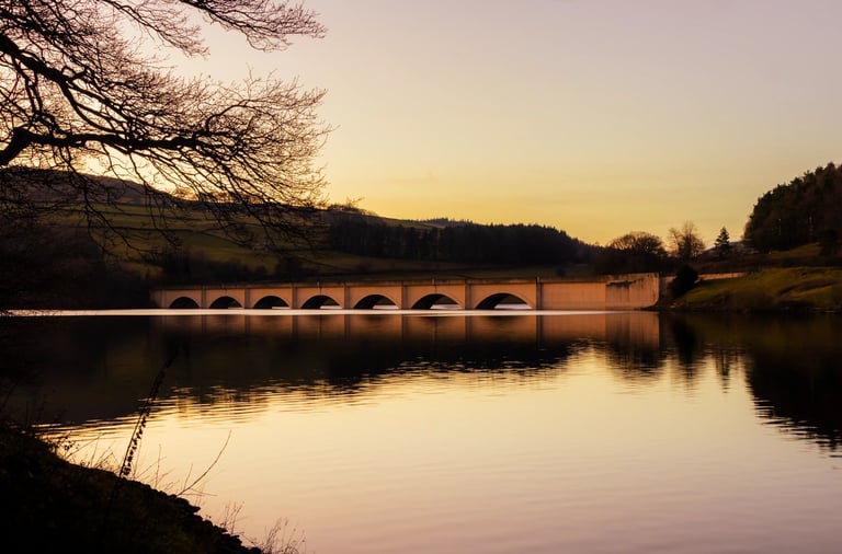 Arched stone bridge reflecting in a calm lake at sunset in a rural landscape.
