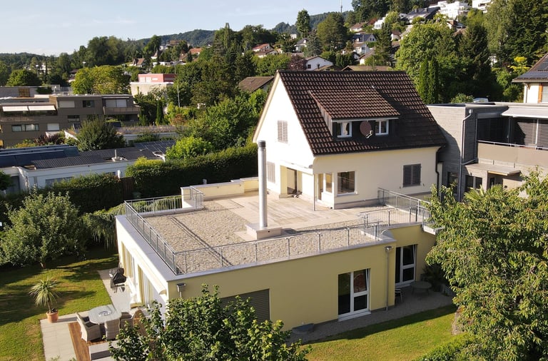 Luftaufnahme eines Vorstadthauses mit großer flacher Dachterrasse und grüner Gartenlandschaft.