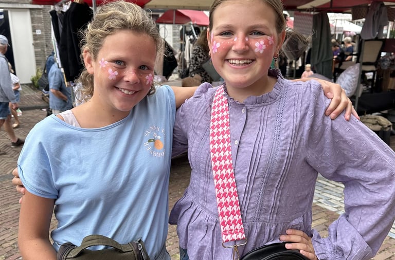 Twee lachende meiden met matchende roze bloemensmink op hun wangen poseren op een markt.