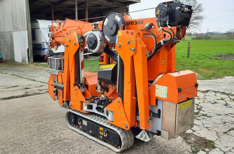 Kompakter oranger Minikran SPD360 auf Raupenketten, zusammengefaltet vor einer Scheune.