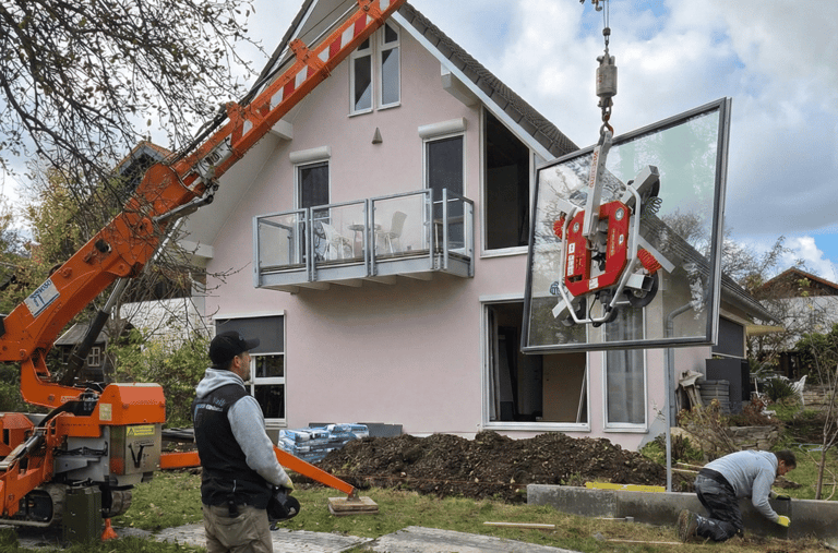 Oranger Minikran hebt mit einem Saugheber eine große Glasscheibe zur Montage in das Obergeschoss eines Wohnhauses.