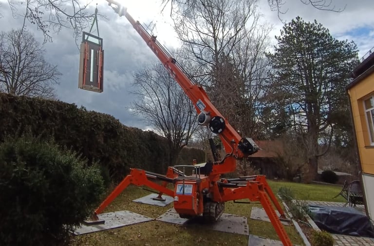 Oranger Minikran hebt ein Bauelement im Garten, stabilisiert durch Stützen auf Bodenschutzplatten.