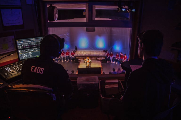 Two figures facing away from the camera looking through a window to the stage