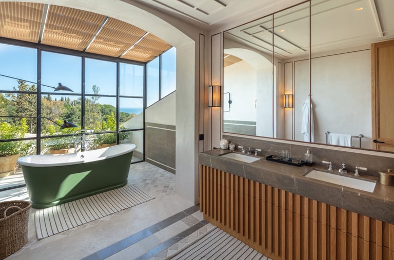 Bathroom with freestanding green tub, double vanity, and floor-to-ceiling garden view windows. 