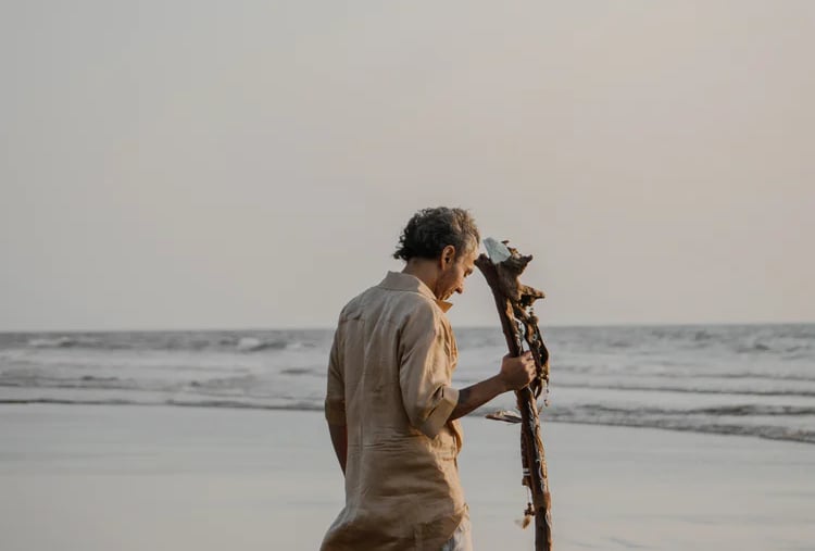 A man in linen clothing walking barefoot on a sandy beach at sunset holding a driftwood staff.