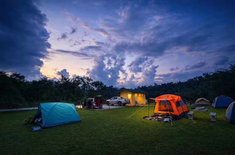 Camping site at dusk with multiple colorful tents, parked vehicles, and a small building under a dramatic, cloud-filled sky.