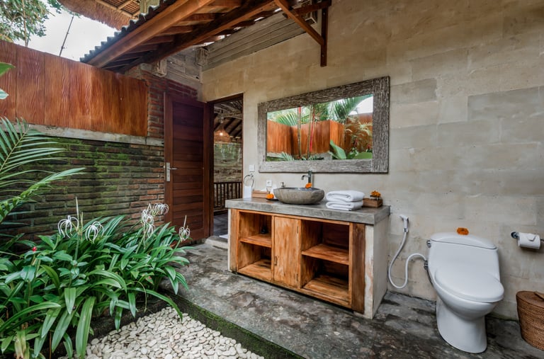 Rustic tropical outdoor bathroom with stone vessel sink, wooden vanity, and white toilet.