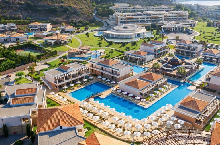 Aerial view of a luxury Greek resort with blue swimming pools, white umbrellas, and elegant villas.